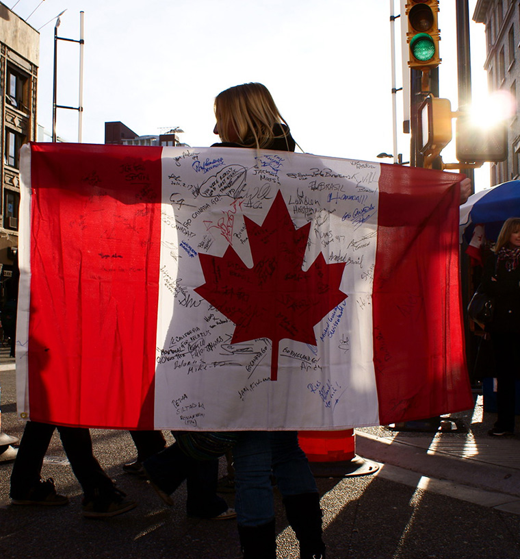 woman facing away has a Canada flag outstretched by both arms across her back, with many signatures and messages handwritten across the flag. A green traffic light and the setting sun appear at top right.